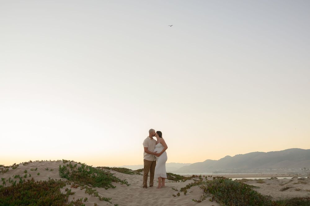 Couple embracing at sunset on sandy dunes with mountain backdrop during golden hour beach engagement session.