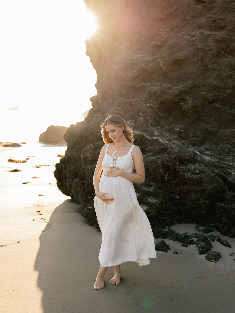 Pregnant woman in white dress on beach at golden hour, cradling baby bump near rocky cliffs.