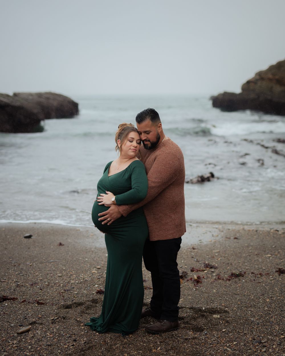 A couple shares an intimate moment on a rocky beach with crashing waves and overcast skies in the background.