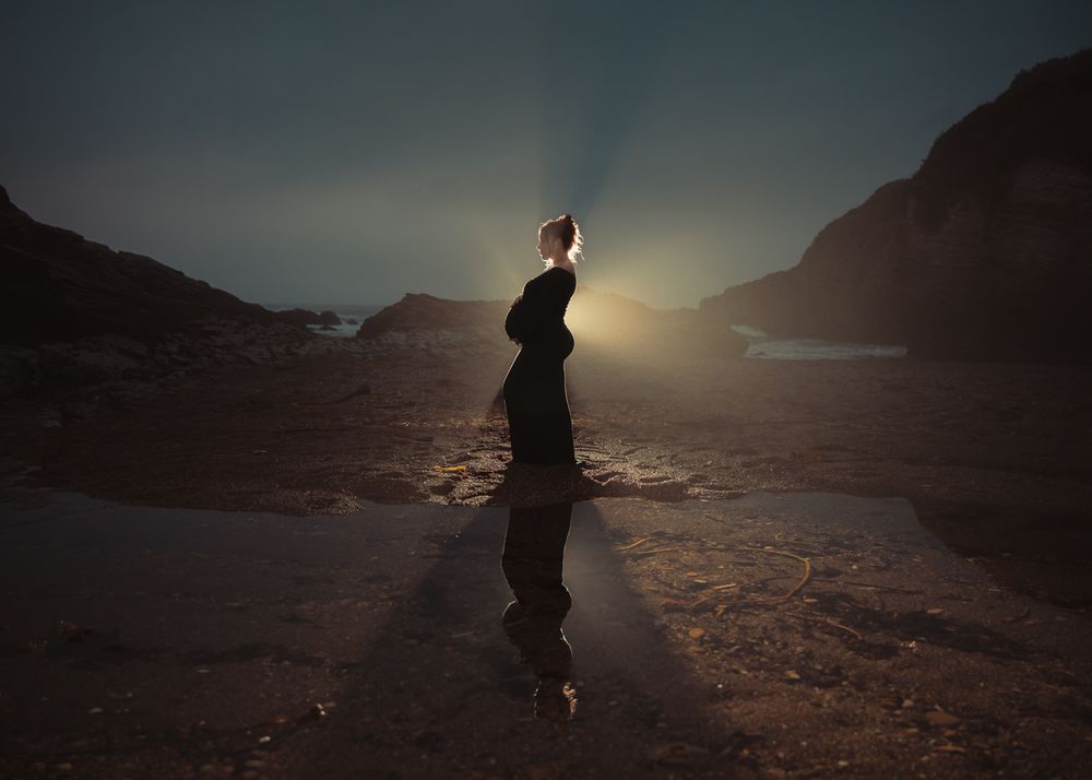A silhouetted figure in a flowing black dress stands on a rocky outcrop against a dramatic sunset sky.