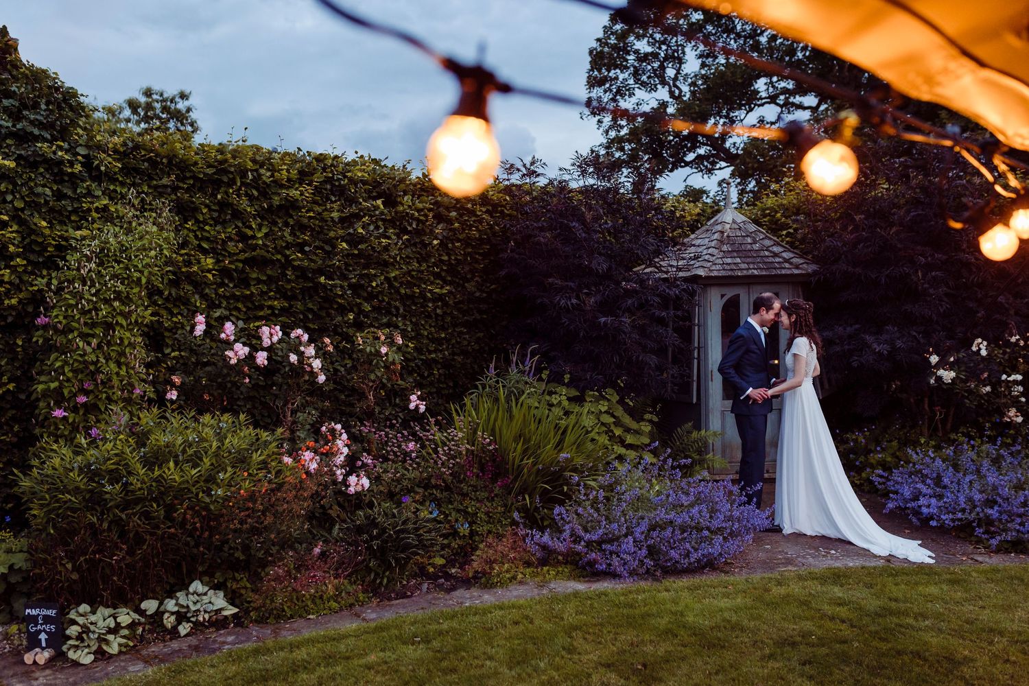 Romantic evening garden scene with string lights illuminating a couple among purple flowers.