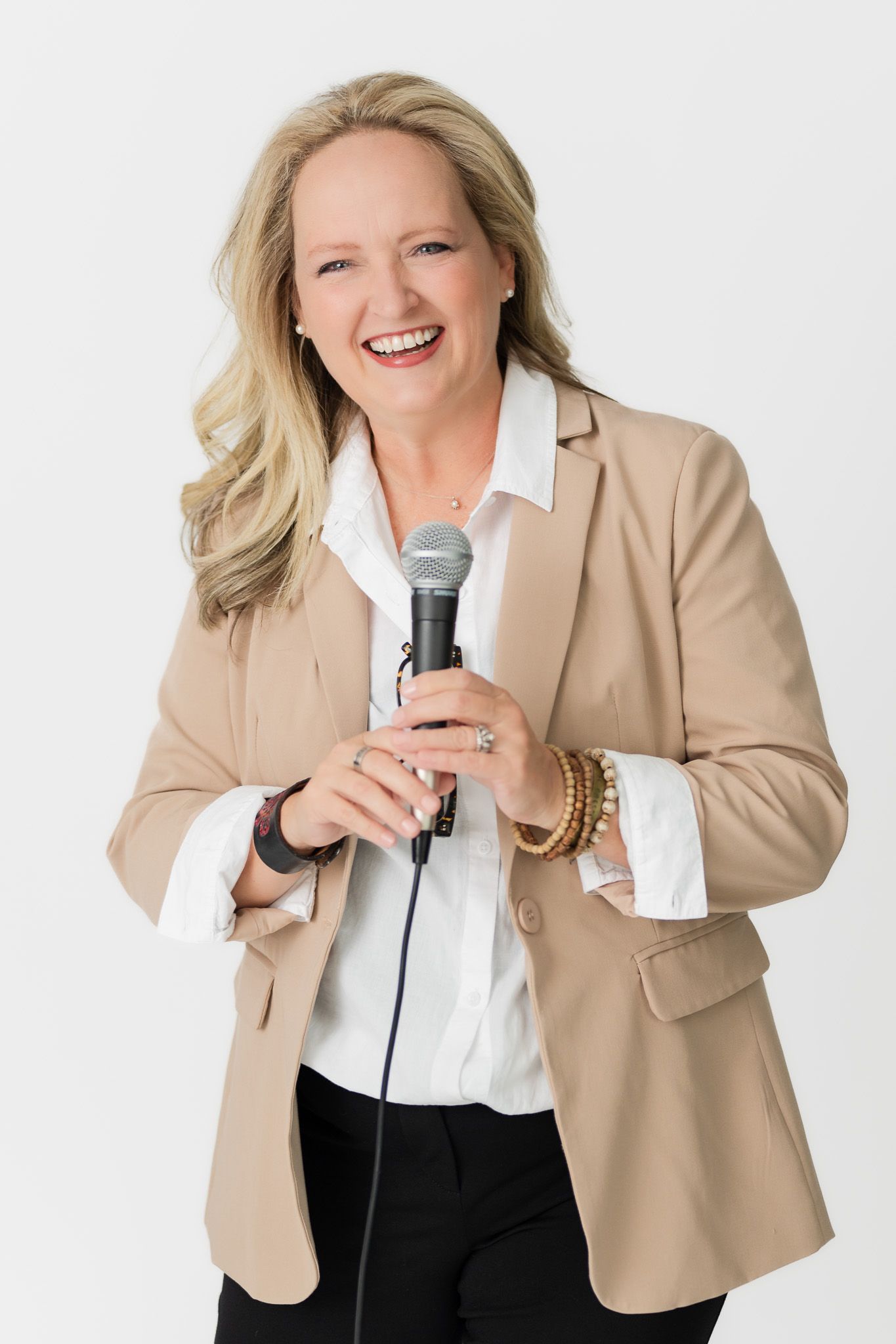Business professional in tan blazer holds microphone while smiling against white backdrop.