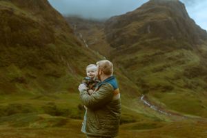 A father holding his smiling young child close as they stand in front of the sweeping, dramatic green mountains of Glencoe, Scotland.