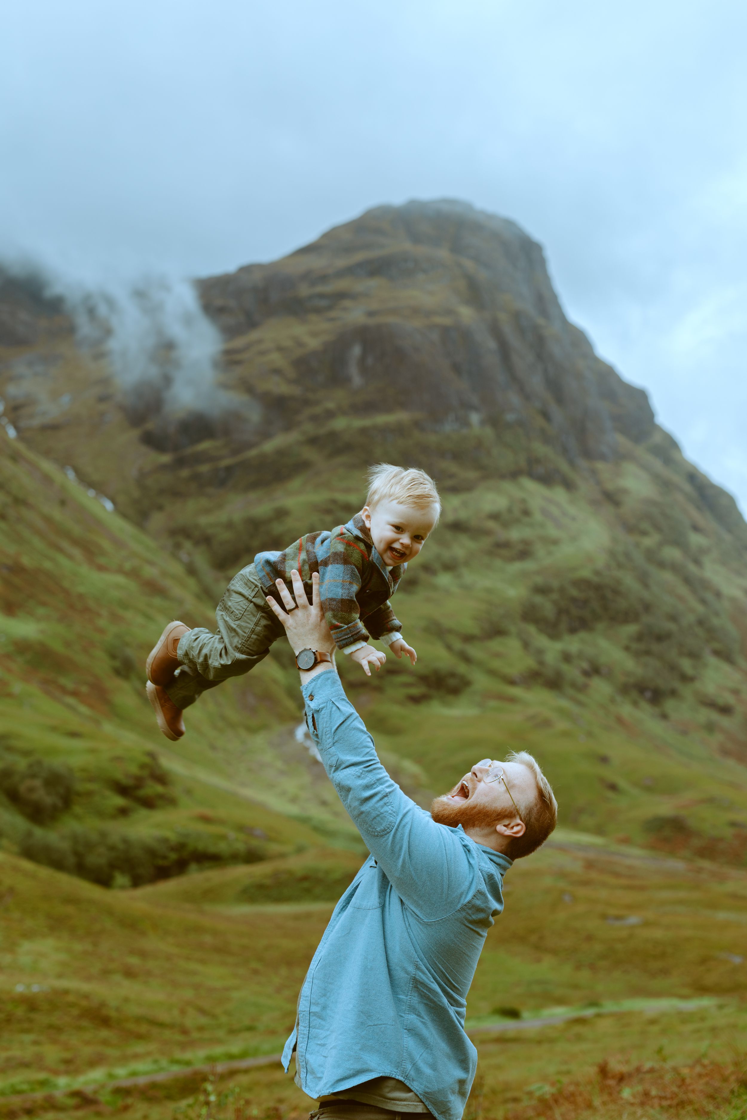 A father holding his smiling young child close as they stand in front of the sweeping, dramatic green mountains of Glencoe, Scotland.