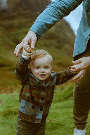 A smiling toddler boy in a plaid jacket taking steps while holding his parent's hands against a green hillside background in Glencoe