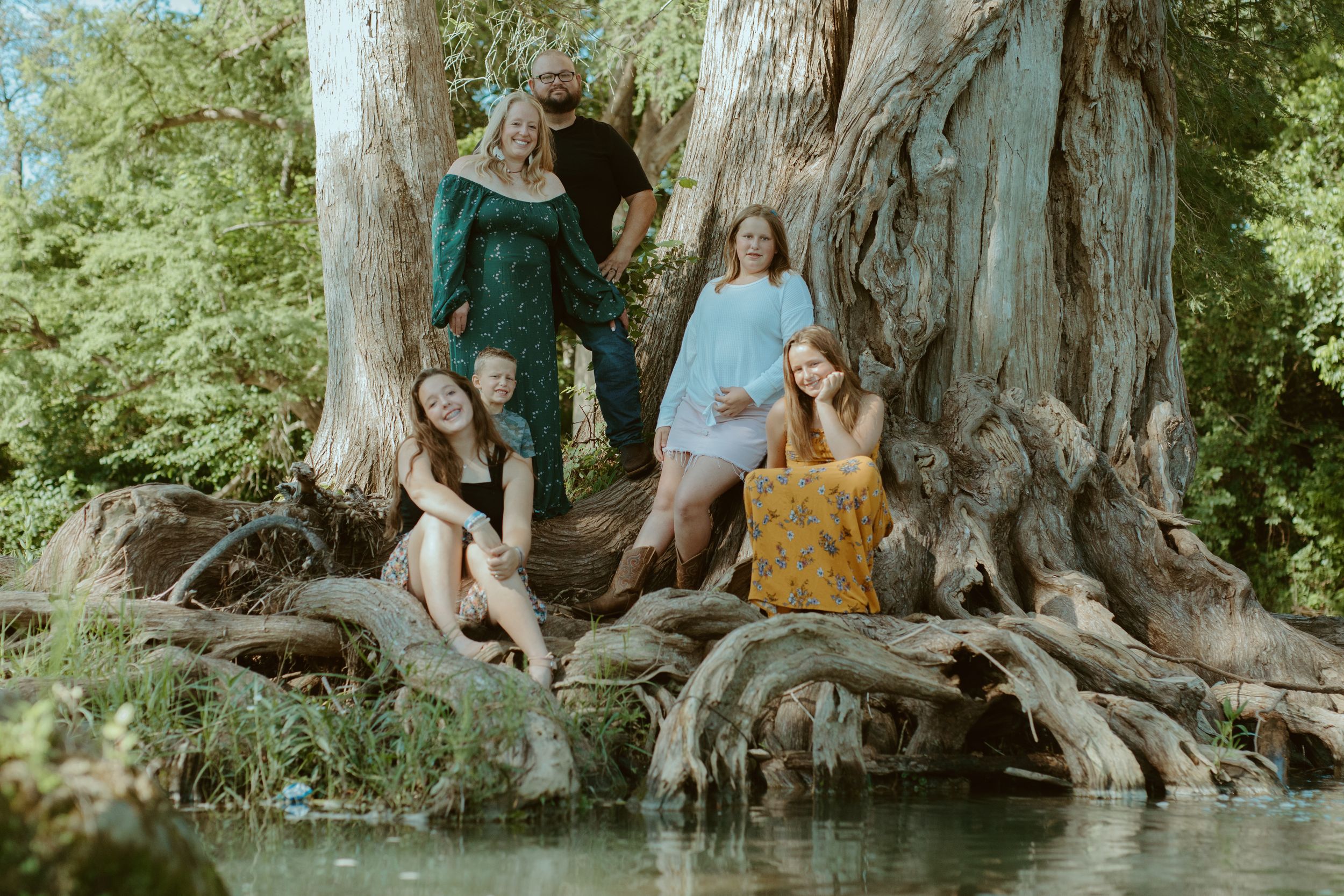 A large family posing together on the massive, exposed roots of a cypress tree along a riverbank in Central Texas.