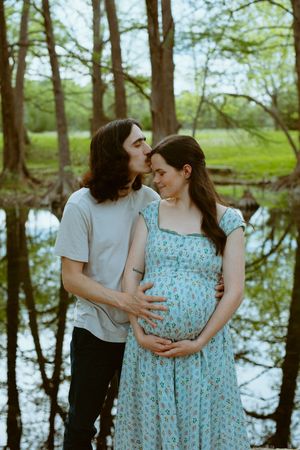 A vertical color photo of a man and a pregnant woman standing on a stone ledge next to a calm river.
