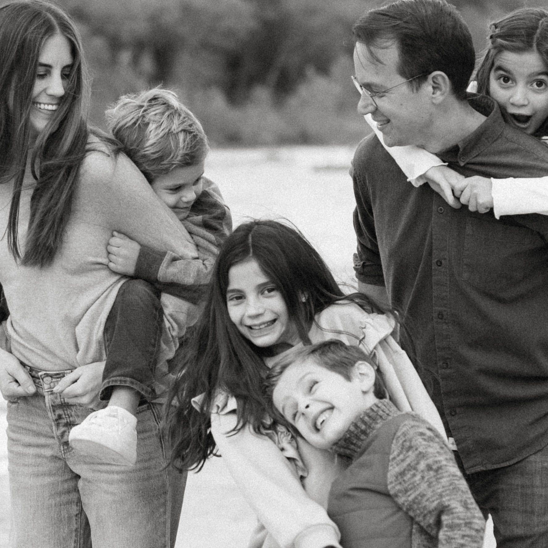 A close-up black and white version of a family photo session by the river, showing parents giving their children piggyback rides while every