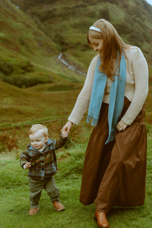 A mother in a blue scarf holding her toddler son's hand as they walk together through the green mountain landscape of Glencoe, Scotland.