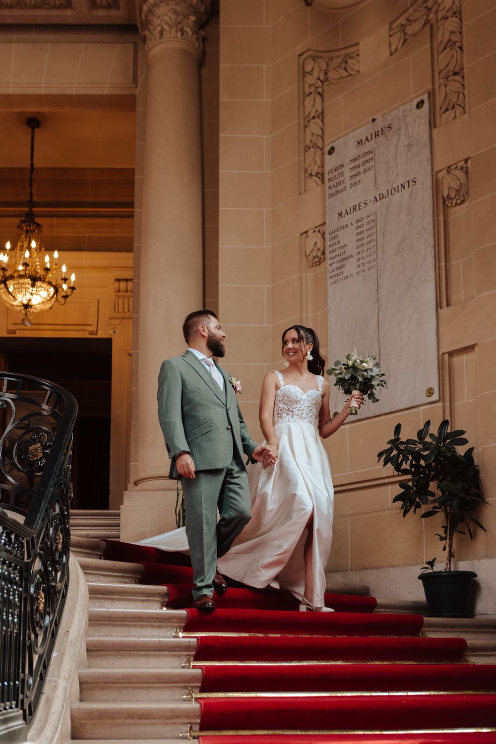 Bride and groom holding hands on red-carpeted stairs in an elegant French wedding venue.