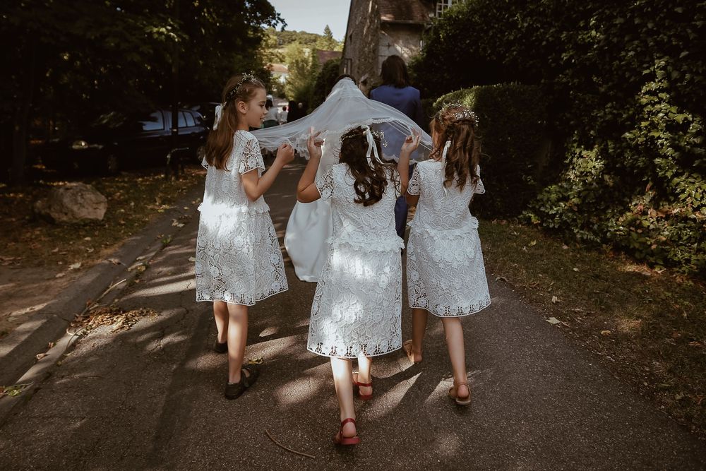 Three flower girls in white lace dresses carry bride's veil down a tree-lined path.