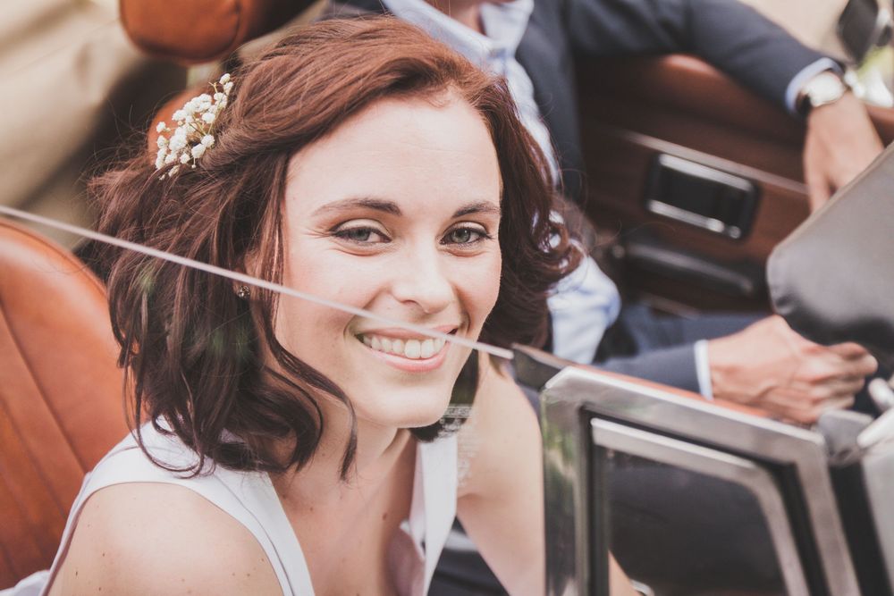 Smiling bride with floral hair accessory riding in a vintage convertible car.