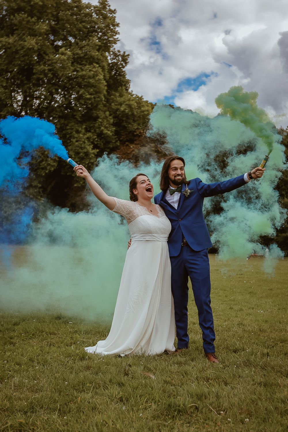 Joyful bride and groom celebrating with colorful blue and green smoke bombs outdoors.