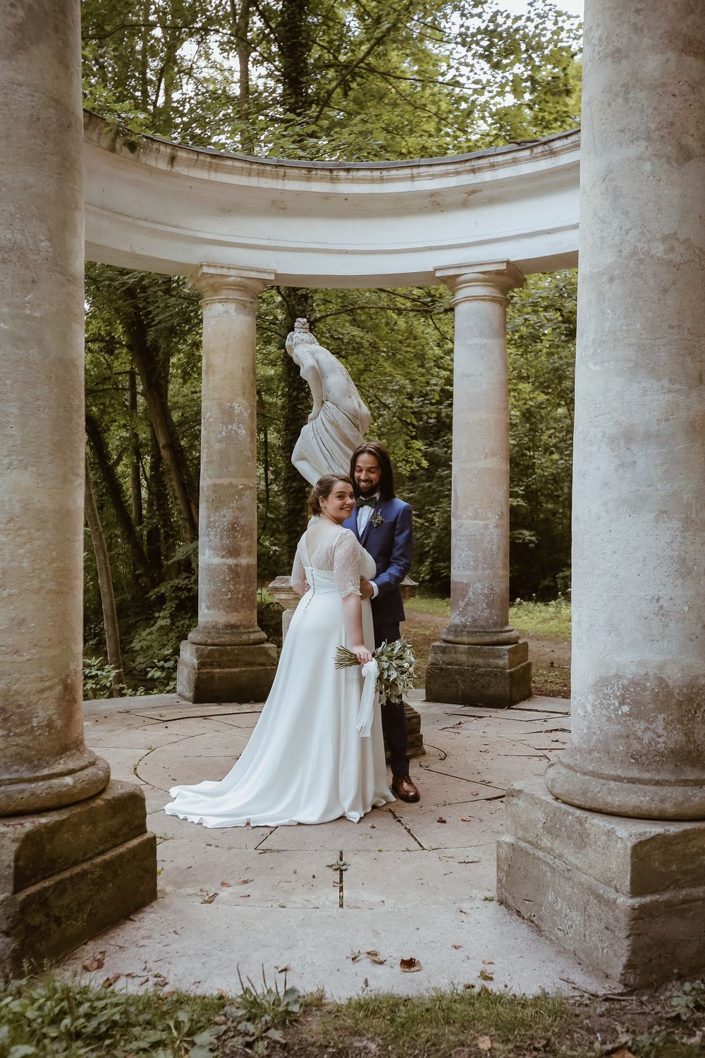 Bride and groom posing among classical stone columns with statue in lush garden setting.