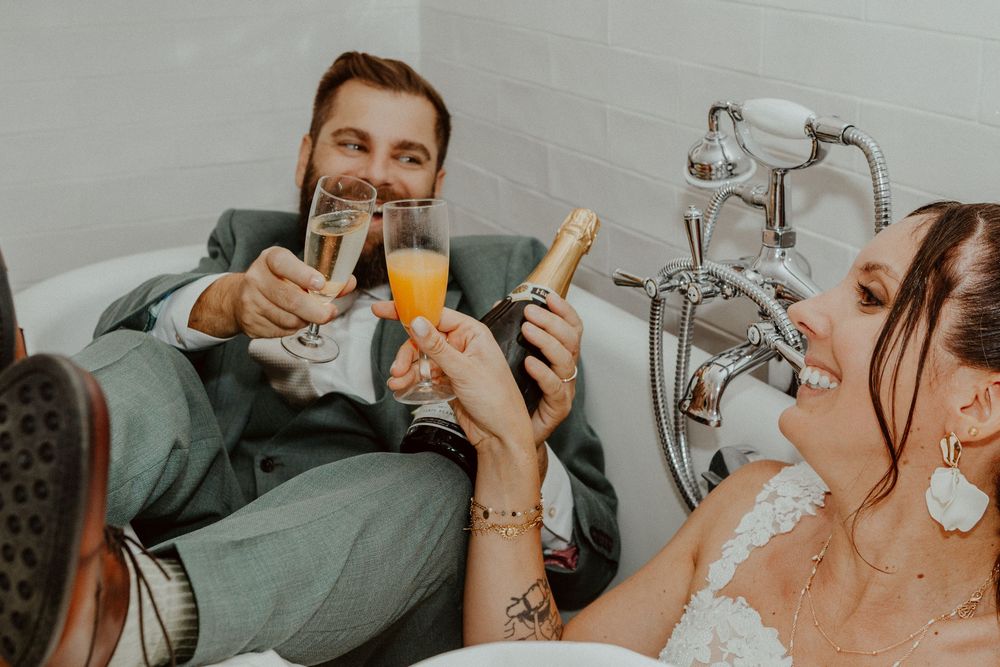 Bride and groom in wedding attire sitting in a bathtub, toasting with champagne glasses and mimosas in a celebratory moment.