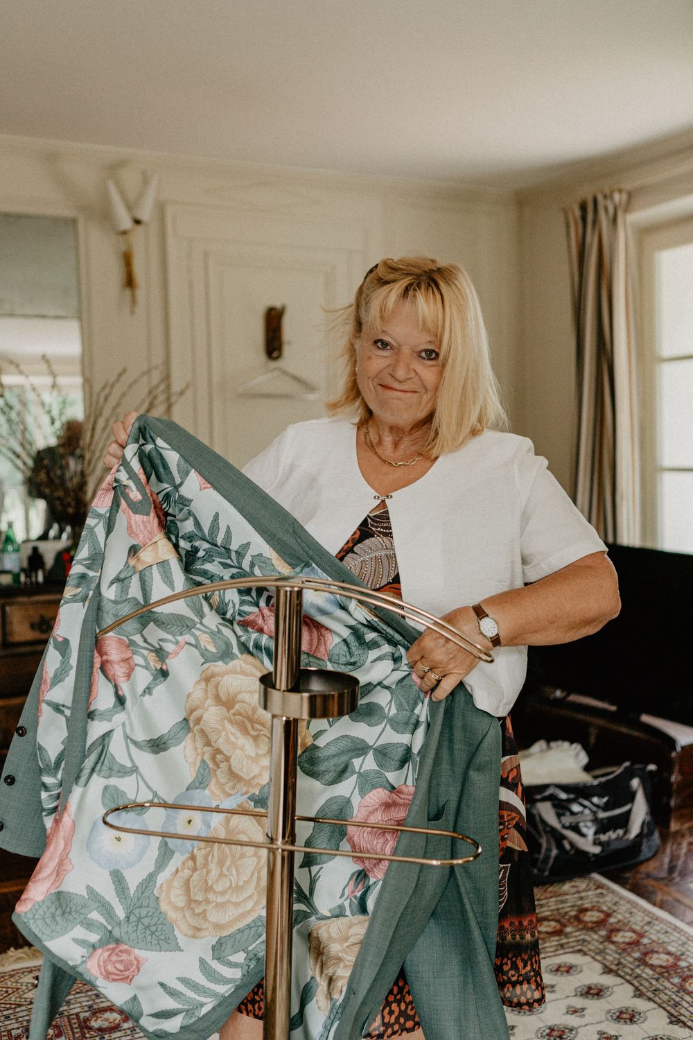 Smiling blonde woman in white jacket holds floral garment on clothing rack indoors.