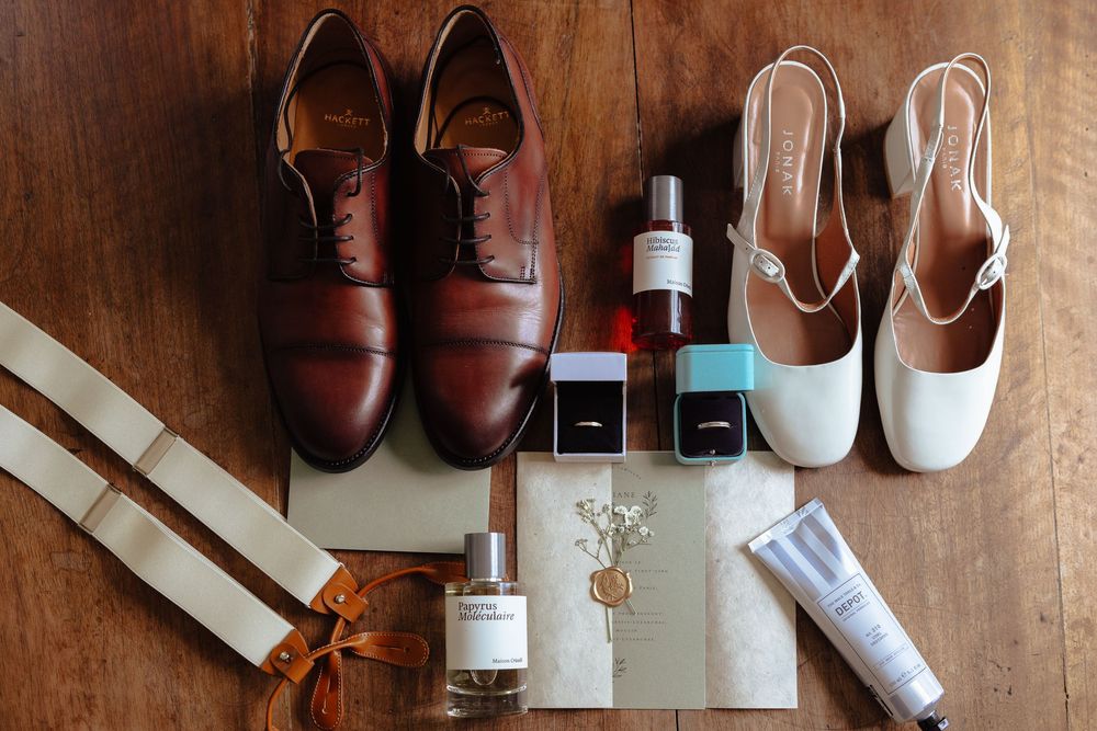 Flat lay of wedding accessories including brown Oxford shoes, white heels, rings, and perfume.