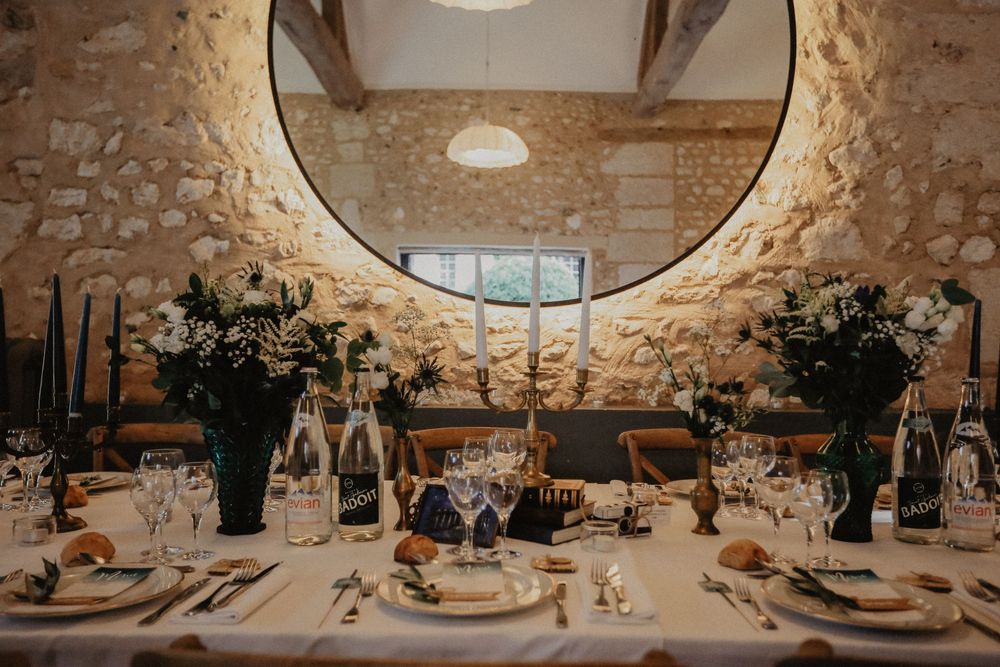 Elegant wedding reception table with floral arrangements, candles, and glassware set against a rustic stone wall with circular mirror.