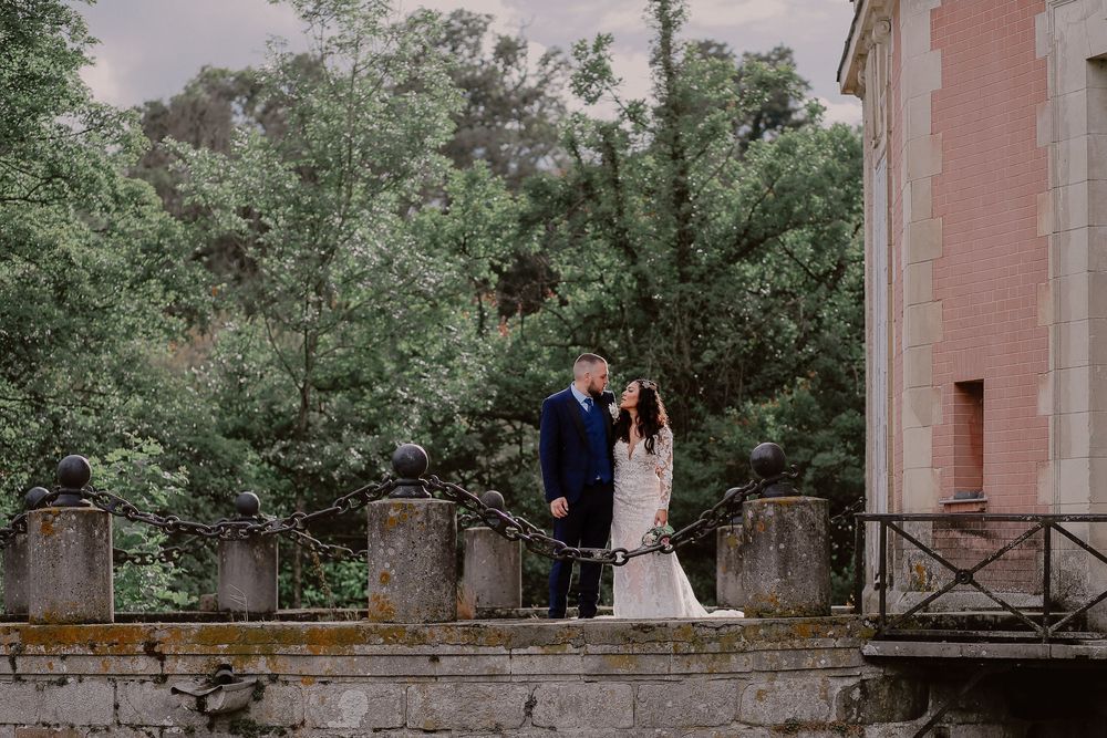 Bride and groom stand together on a stone terrace surrounded by lush green trees at an elegant outdoor wedding venue.