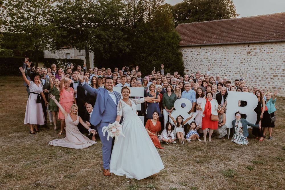 Large wedding group photo with bride and groom posing outdoors, guests holding decorative letters at a countryside venue.