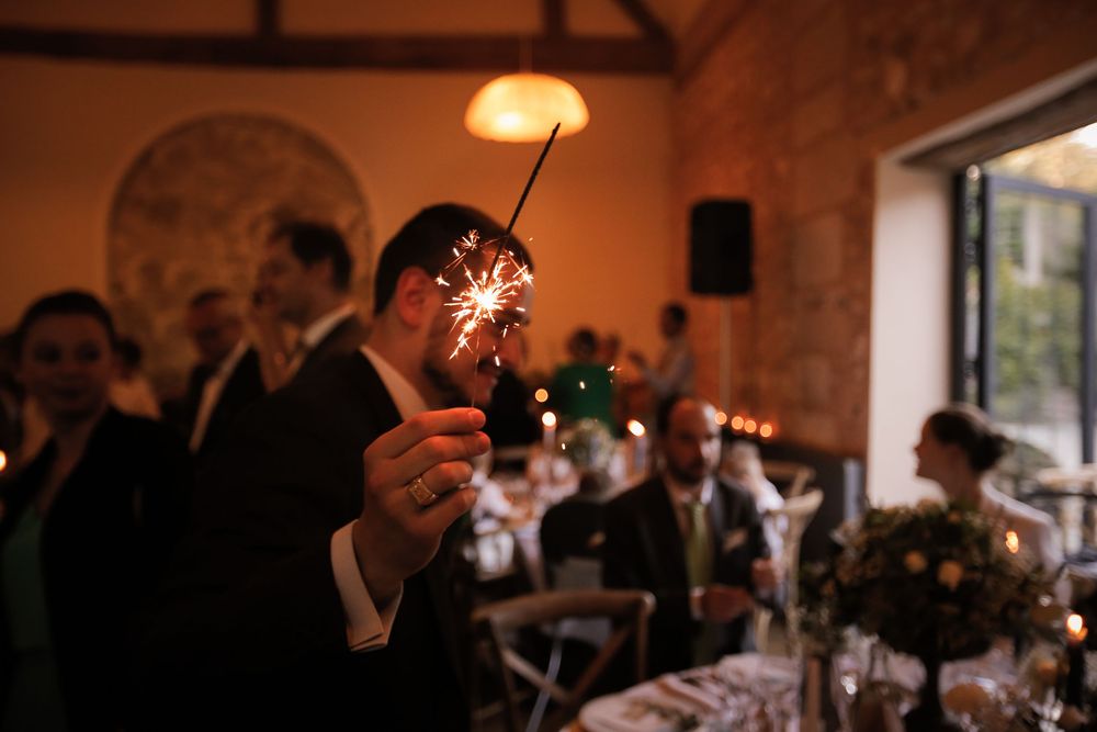 A wedding guest holds a sparkler at an elegant indoor reception, with guests and floral centerpieces visible in the warm background.
