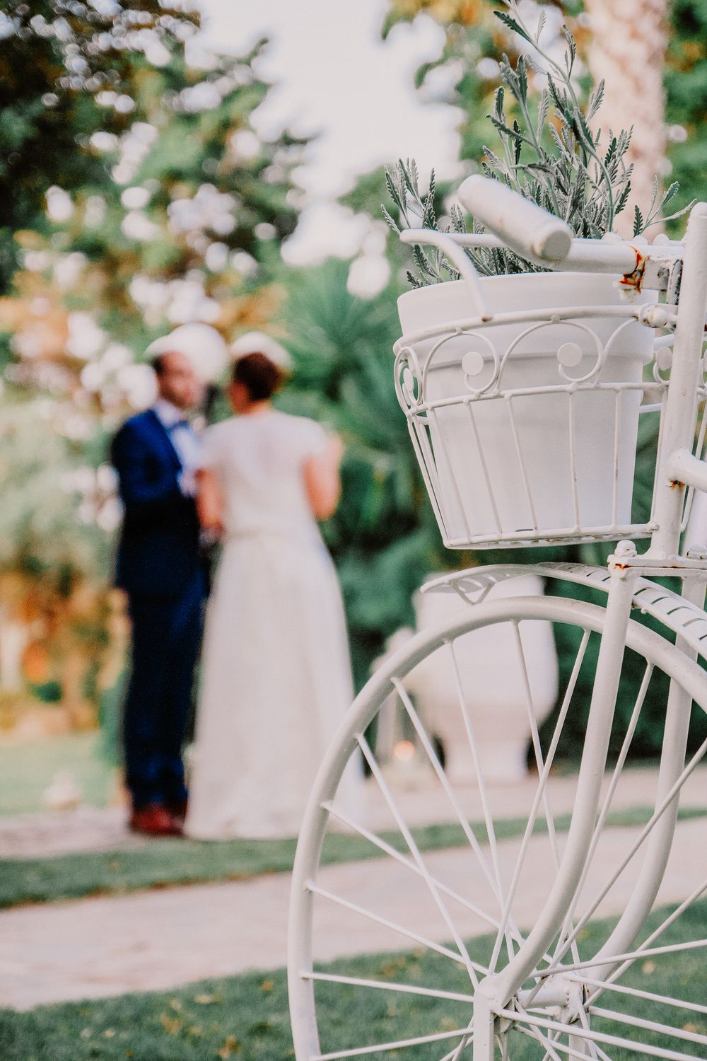 White decorative bicycle with basket in foreground, blurred bride and groom embracing in lush garden background.