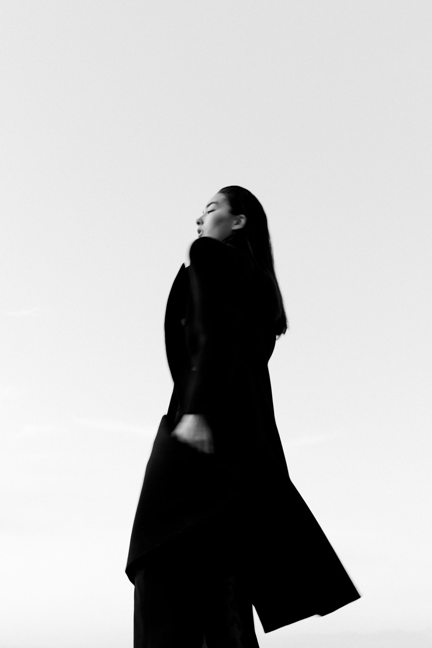 Artistic black and white portrait of woman in flowing coat against white background.