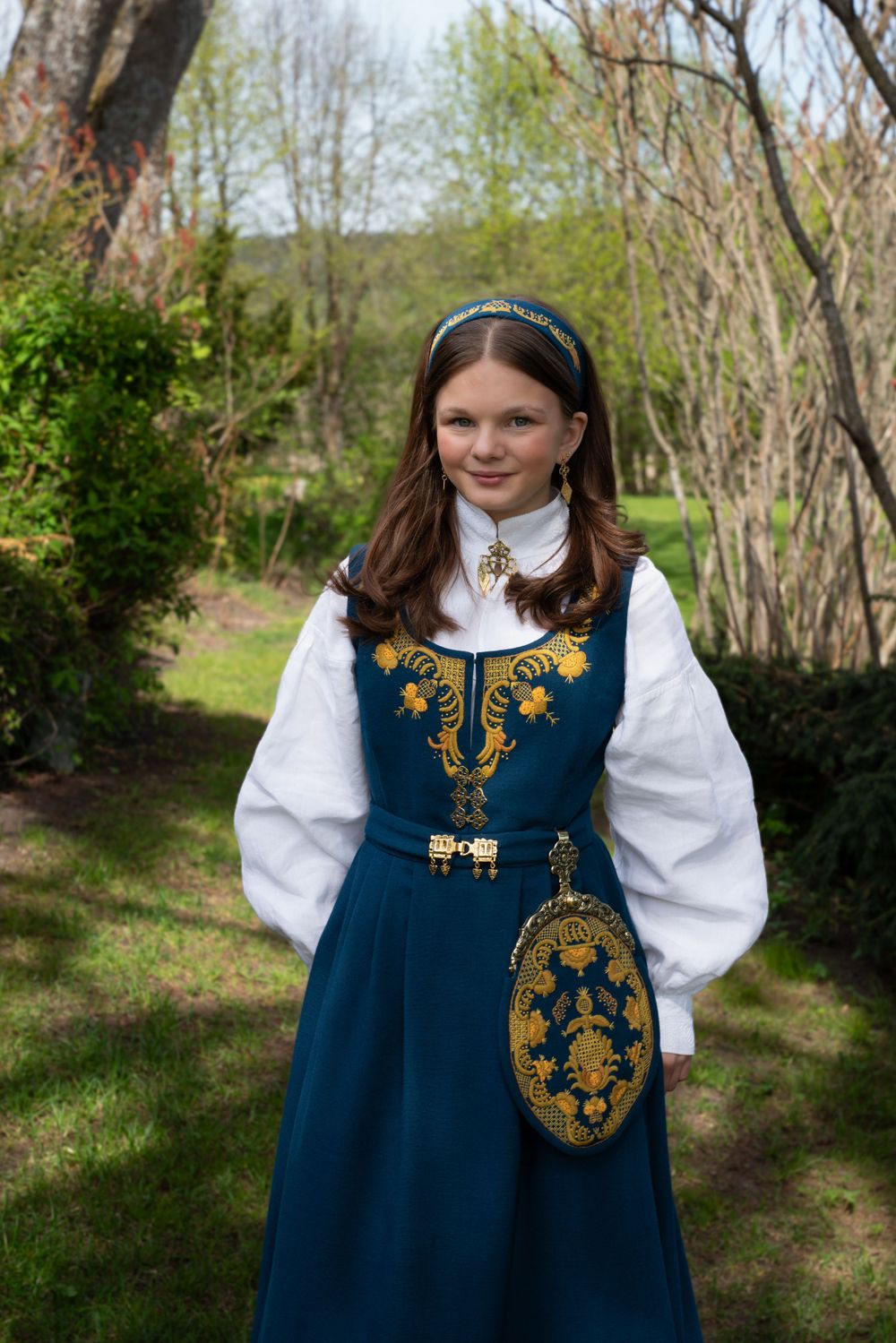 Young person in traditional Norwegian bunad dress with embroidered details and matching purse in garden setting.