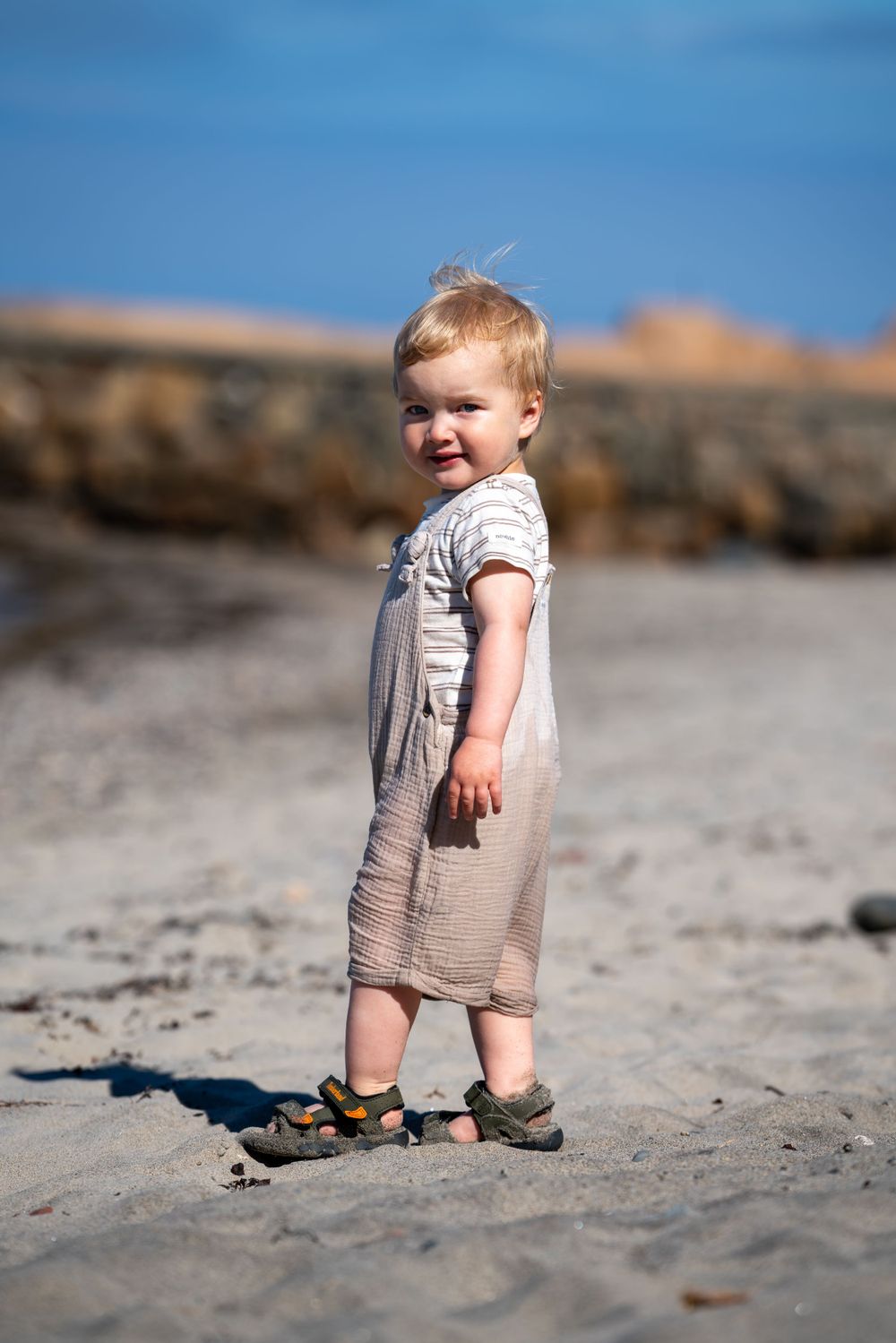 Small child standing on sandy beach wearing light colored overalls on a sunny day with rocks in background.