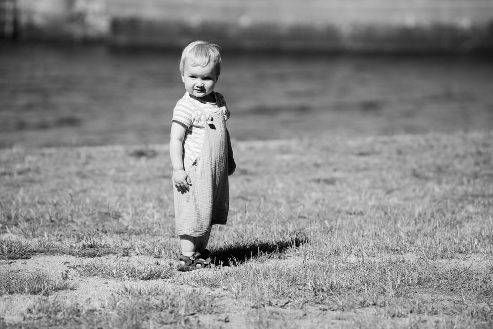 Vintage black and white photo of toddler standing alone in grass wearing light dress.