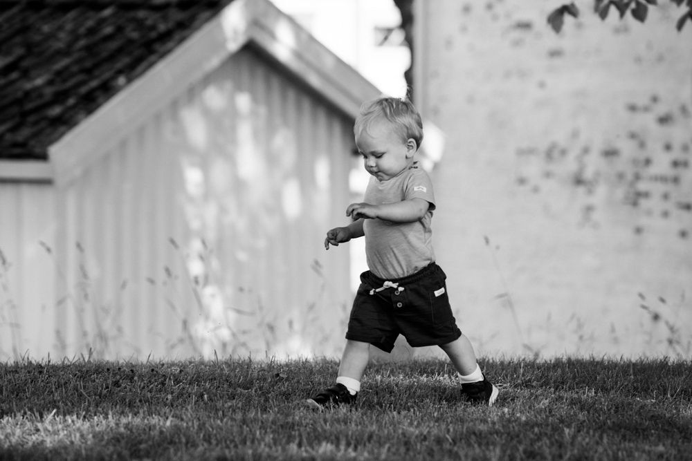A toddler takes steps on grass near white buildings on a sunny day.