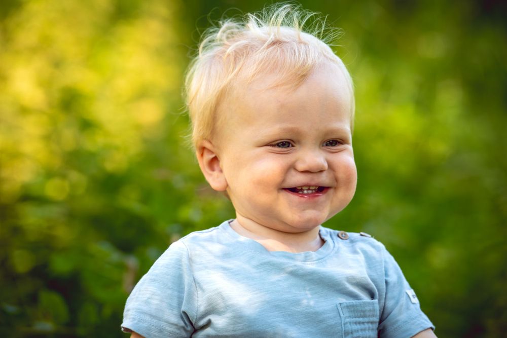 A cheerful toddler smiles outdoors against a blurred green background.