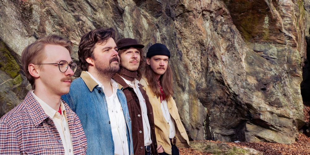 Four young men standing outdoors against a rocky cliff face, dressed in casual vintage-style clothing.