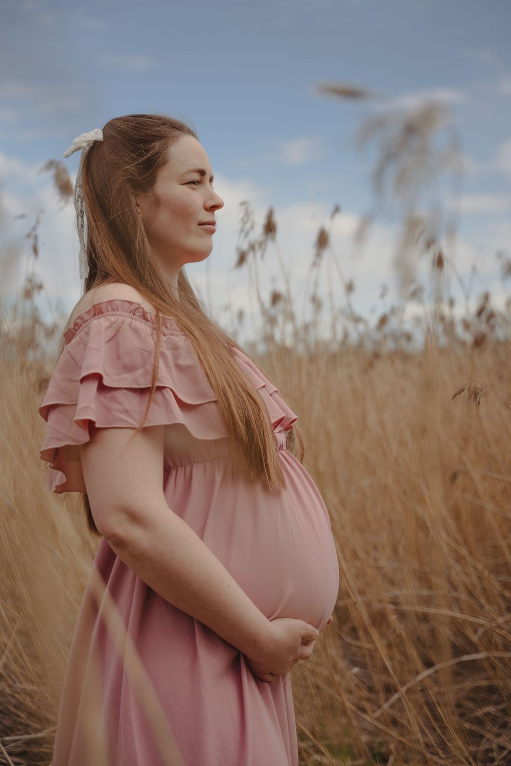 Woman wearing pink ruffle dress poses thoughtfully in tall autumn wheat field during golden hour.