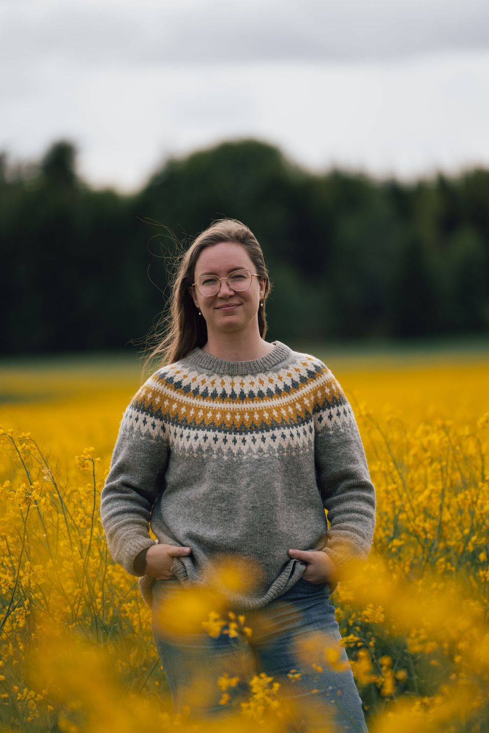 Someone wearing a gray and yellow Fair Isle sweater poses playfully in a vibrant yellow canola field on an overcast day.
