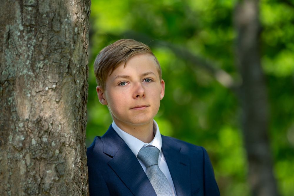 Young boy in navy suit and gray tie posing beside a tree in a lush green forest setting.
