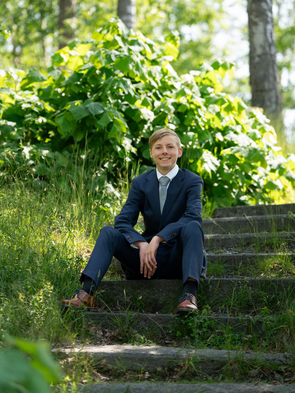 Young man in navy suit sitting on stone steps surrounded by lush green foliage outdoors.
