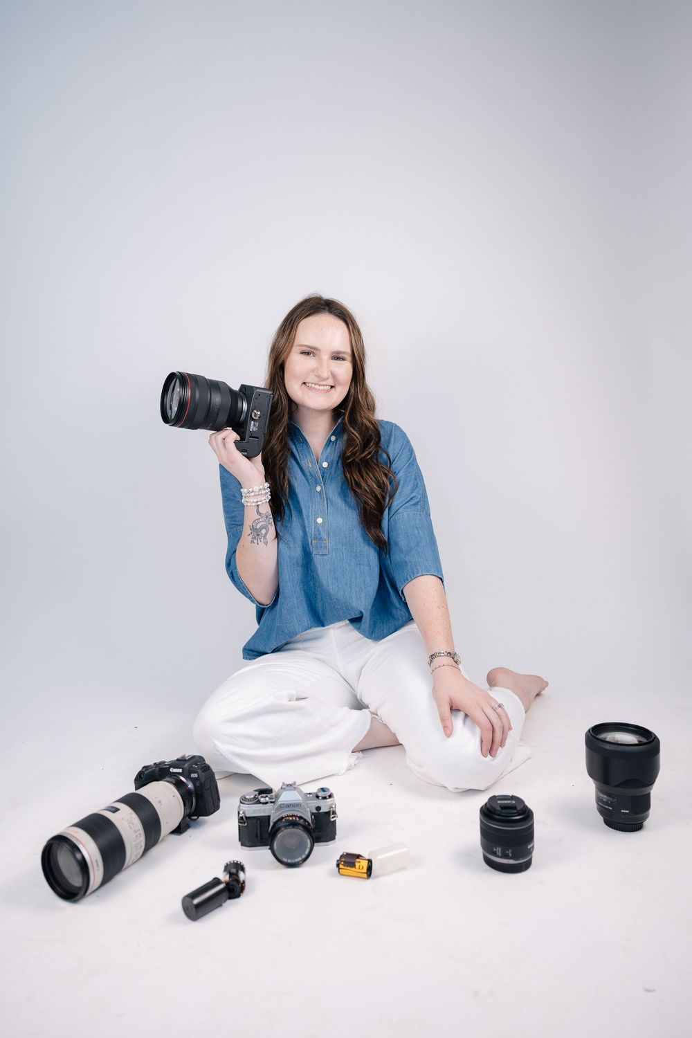 Photographer in blue denim shirt displays camera equipment on white background.