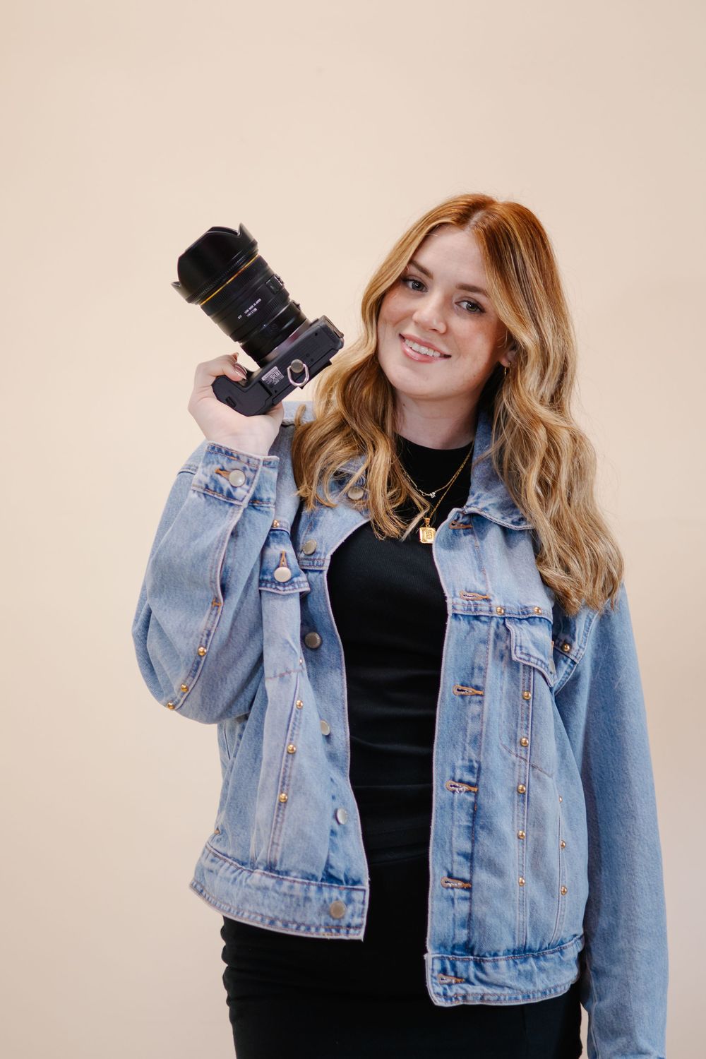 Person wearing denim jacket and black top posing against neutral backdrop.