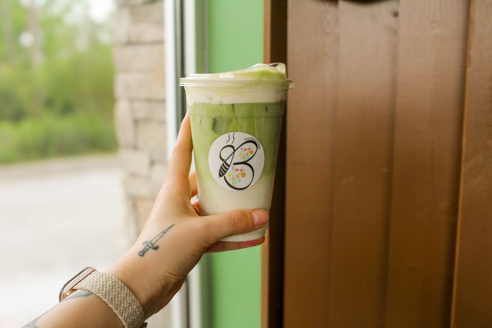 Hand with small tattoo holds a green smoothie drink in a clear plastic cup with a green logo against wooden backdrop.
