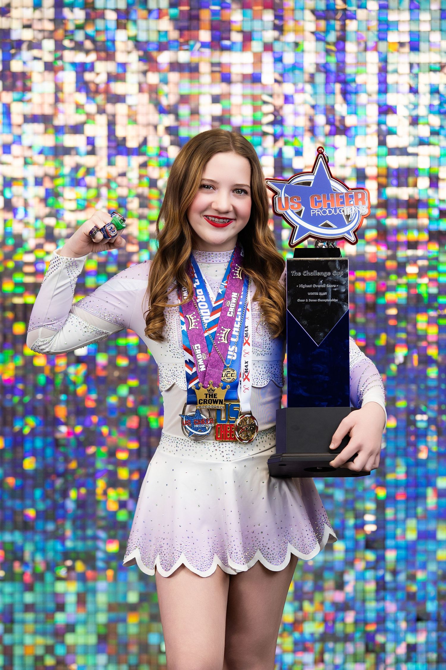 Figure skater in white dress holds trophy and medals against sparkly blue mosaic background.
