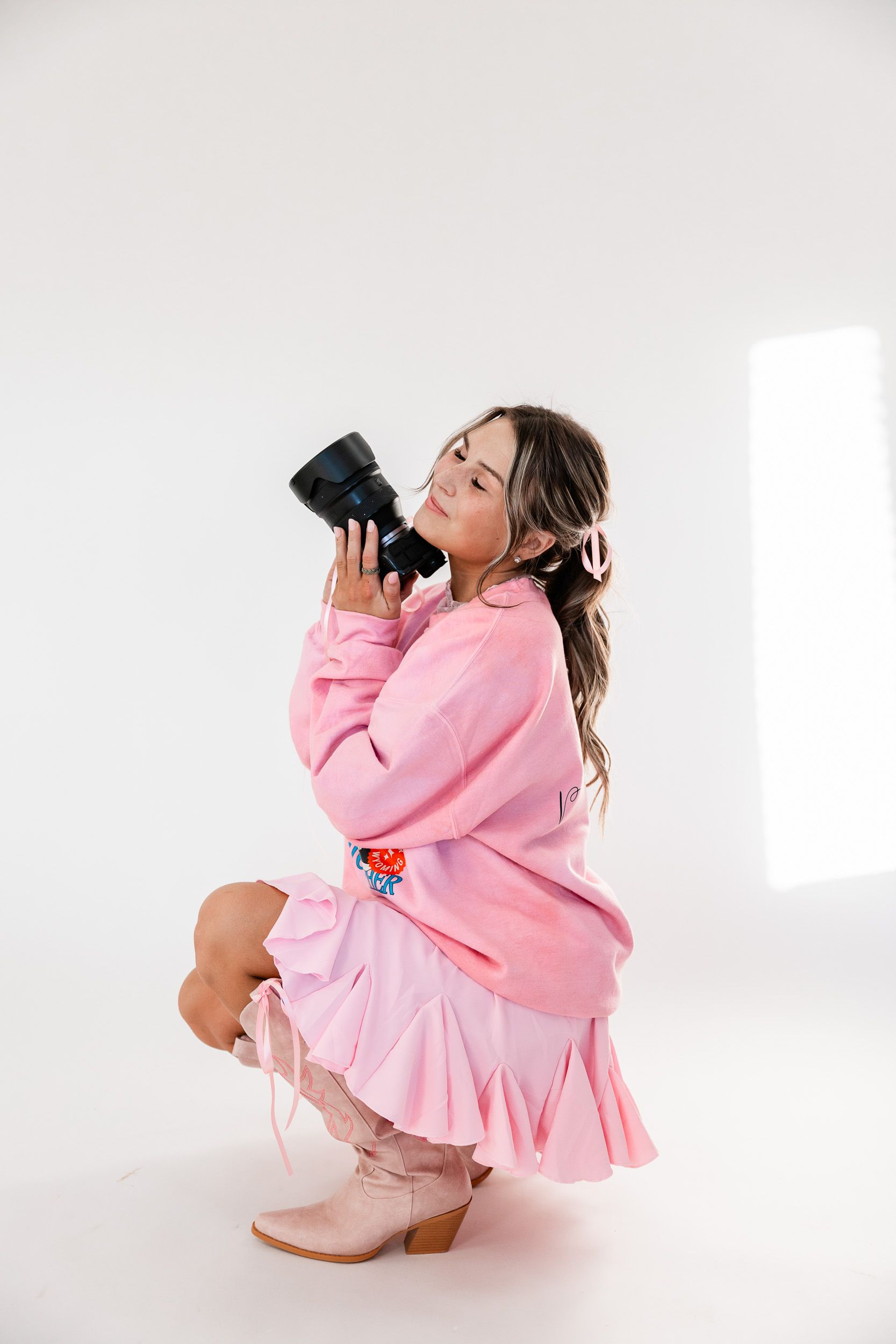 Wyoming Photographer
Person wearing pink sweater and ruffled skirt crouches down with camera against light background.