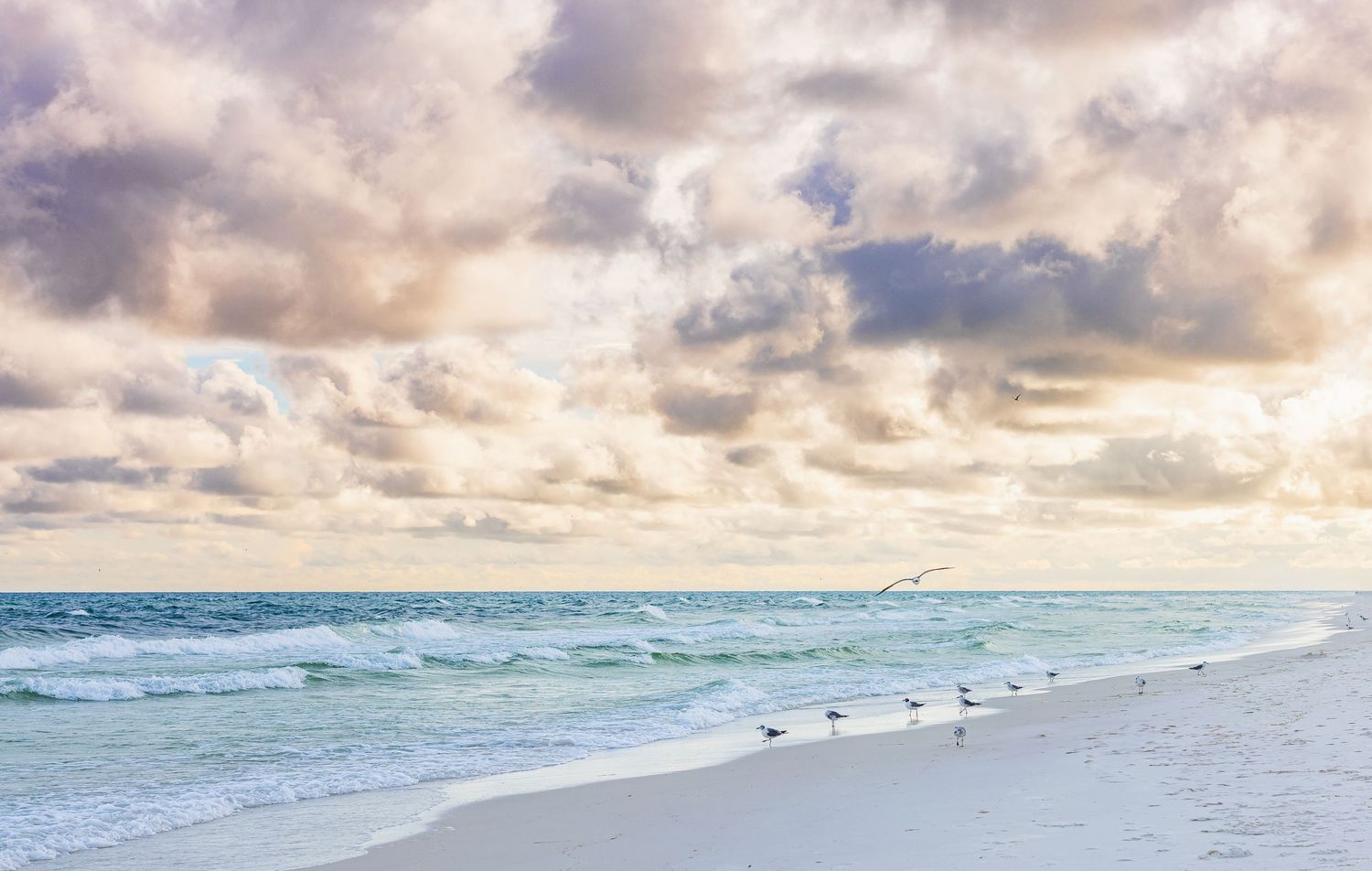 A panoramic beach scene with dramatic cloudy sky and turquoise waves rolling onto white sandy shore at sunset.