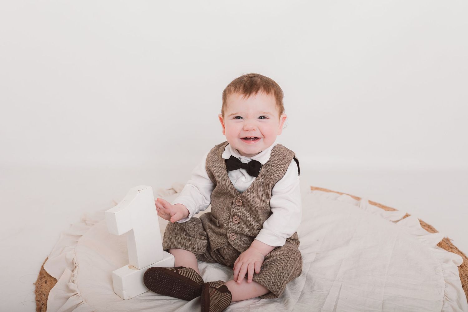 Baby wearing brown overalls and bowtie sitting on cream colored blanket.