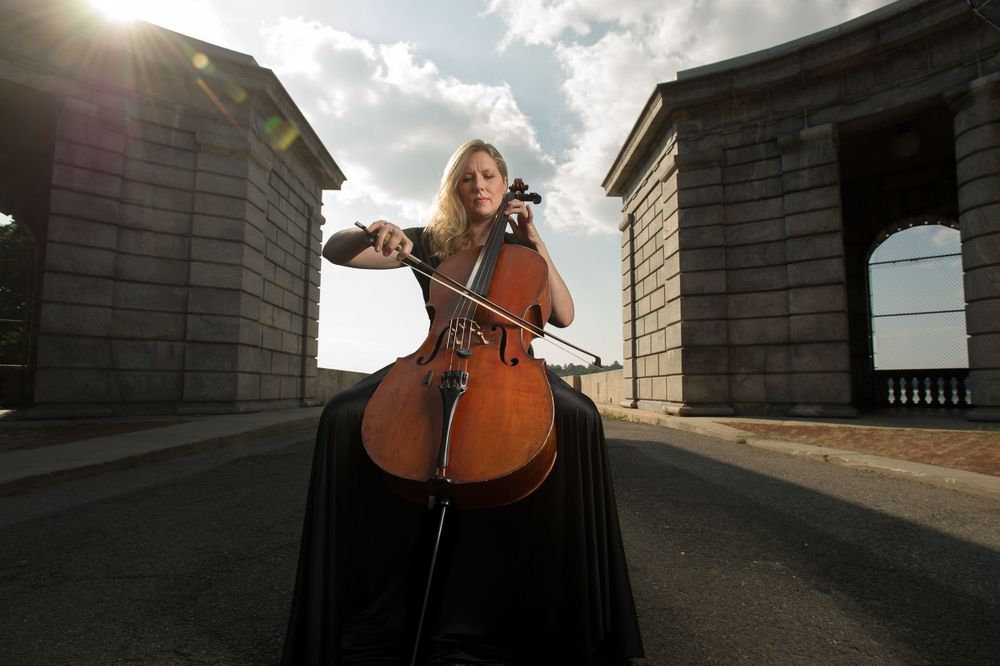 Cellist playing outside in a brick structure with the sun and fluffy clouds in a blue sky