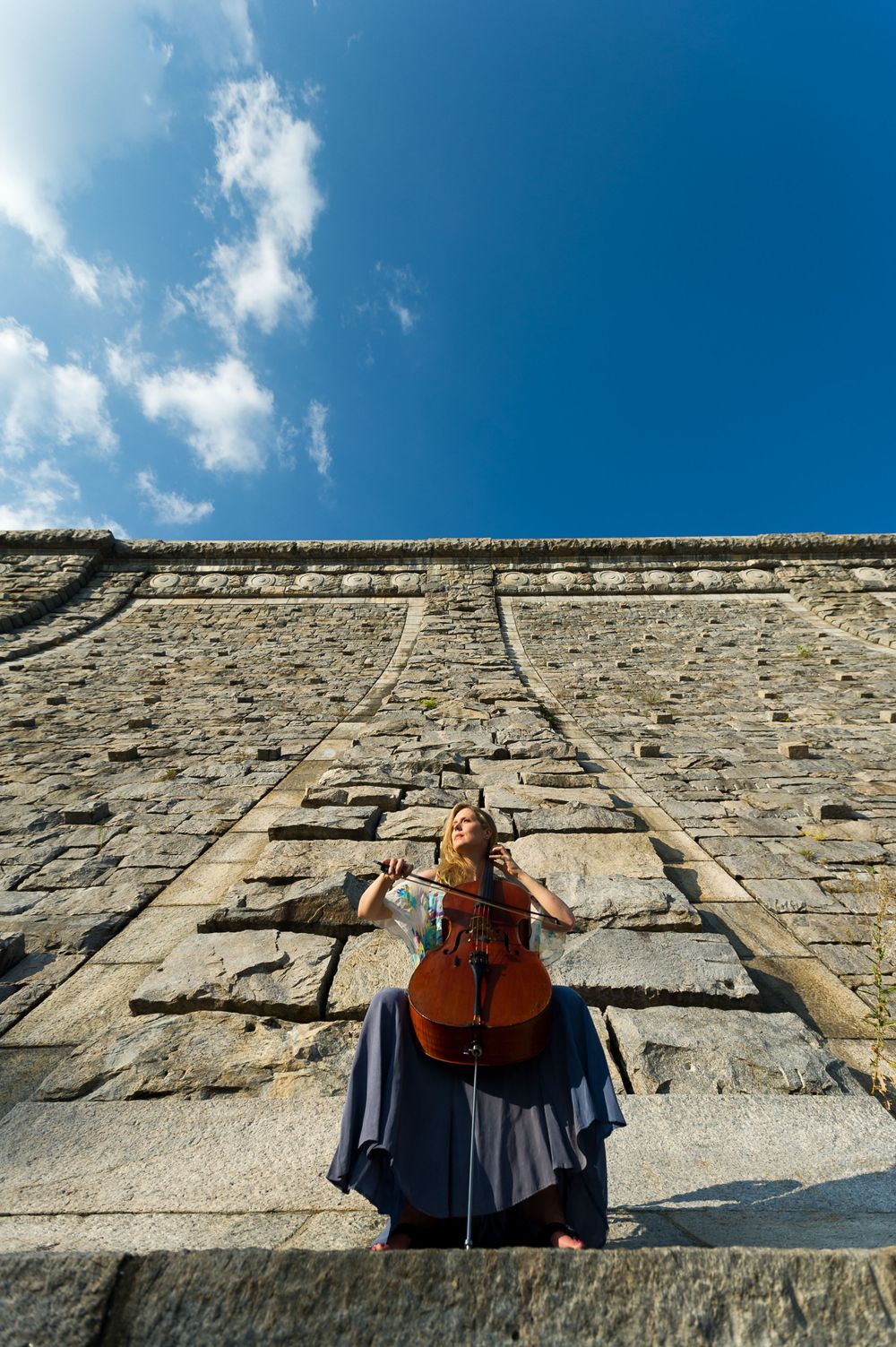 Cellist playing at the base of a stone wall with a bright blue sky and puffy clouds