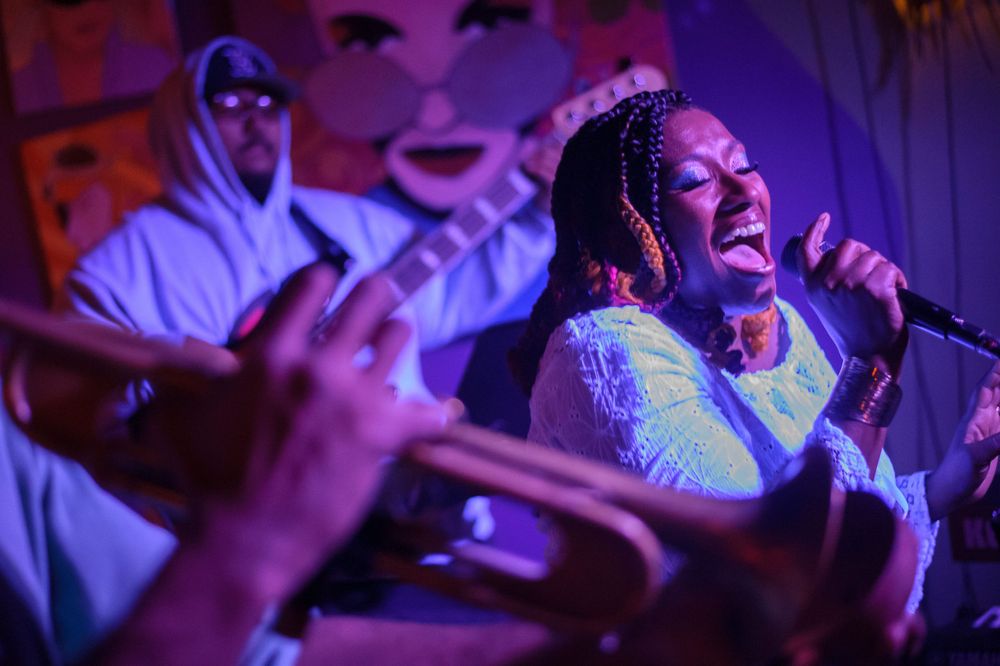 African American female lead singer, framed by trumpet in the foreground and bass player behind, singing into the mic lit by stage lights