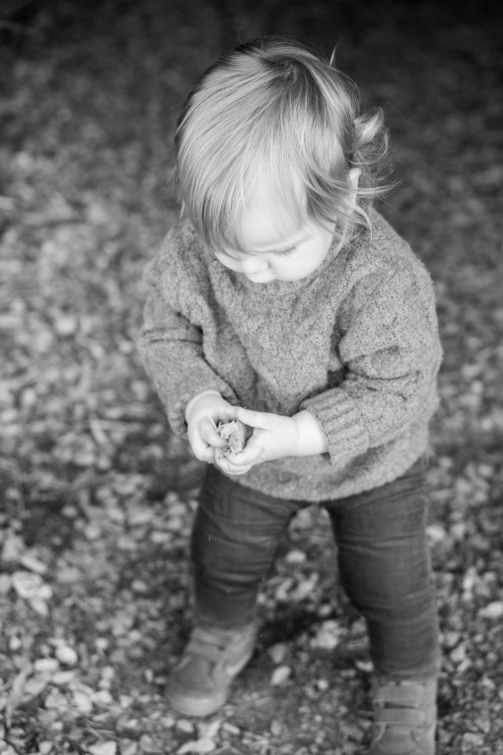 Child in grey sweater and jeans looking down at something in their hands outdoors.