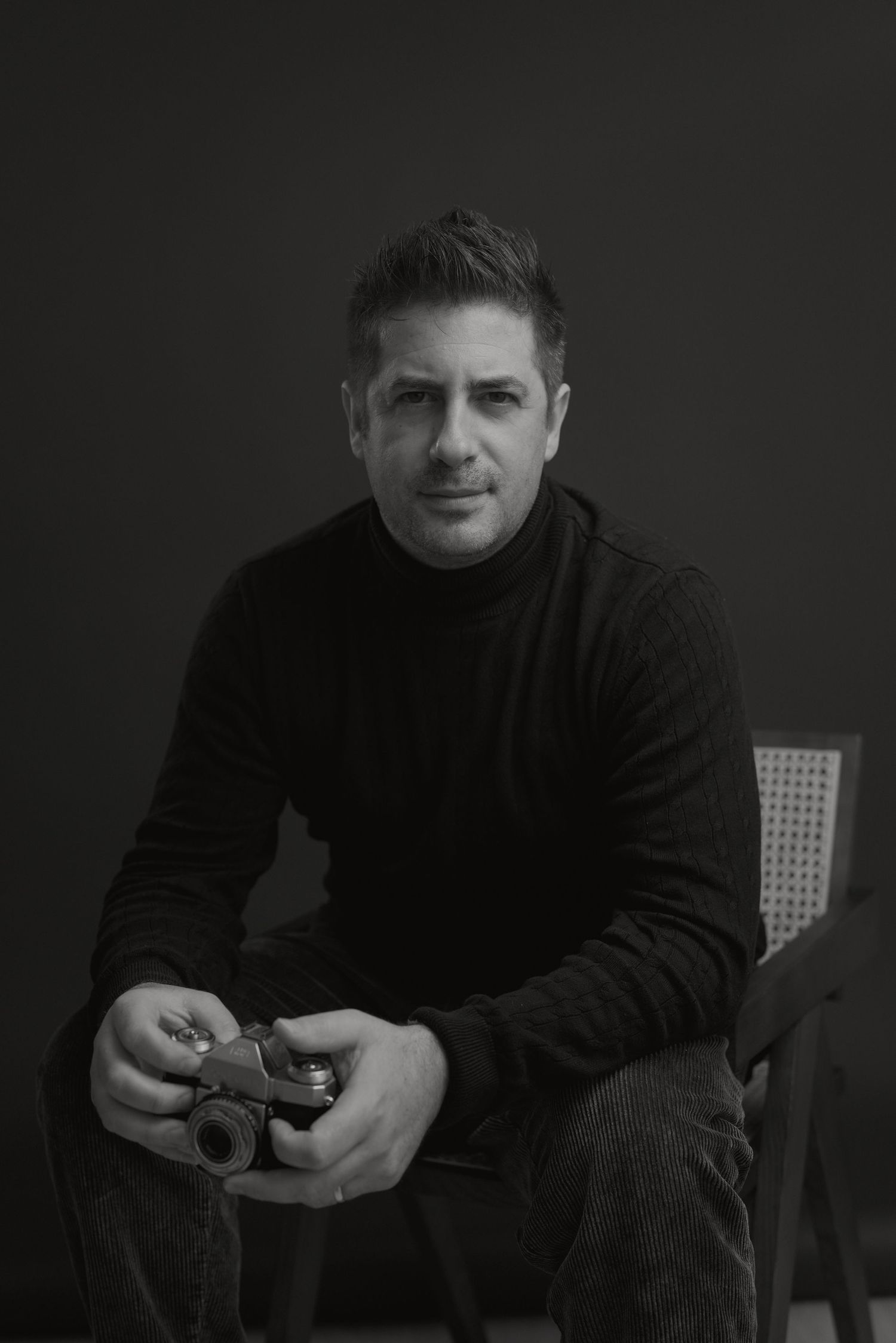 A black and white portrait of someone in a dark turtleneck sweater holding a camera while seated in a chair.