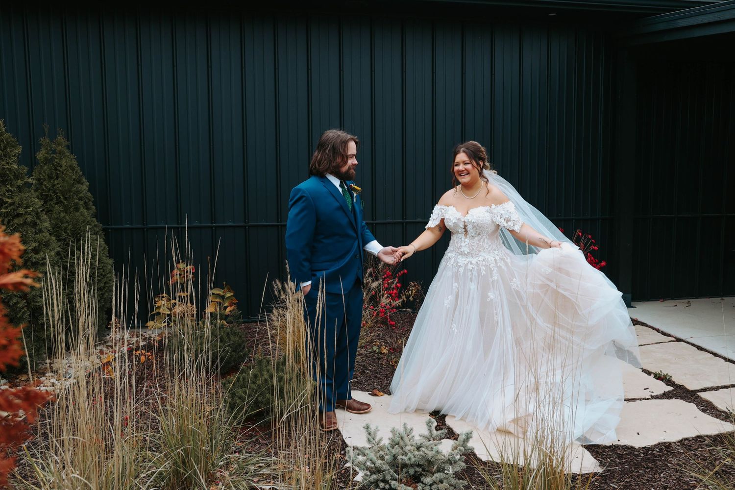 Newlyweds share romantic moment outdoors near dried ornamental grasses against dark building exterior.